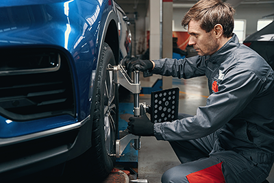 A mechanic in a grey uniform attaching a professional alignment sensor to the wheel of a blue car in a service garage.