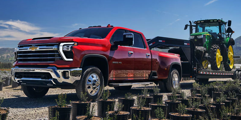 A red Chevrolet Silverado 3500 HD dually towing a gooseneck trailer with a green John Deere tractor through a plant nursery.