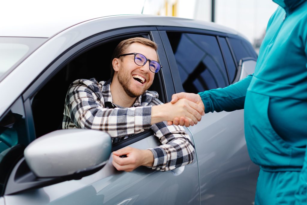 A smiling man sitting in the driver's seat of a silver car shaking hands with a person standing outside the vehicle.