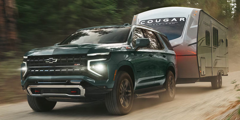 Front-three-quarter view of a dark green Chevrolet Tahoe Z71 driving down a dusty dirt road while towing a large white Cougar Half-Ton travel trailer.