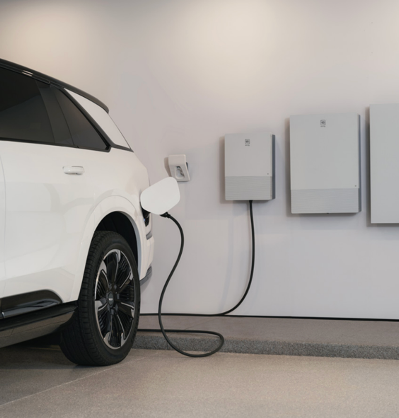 A white electric car is charging through a wall-mounted outlet, with two large batteries below.