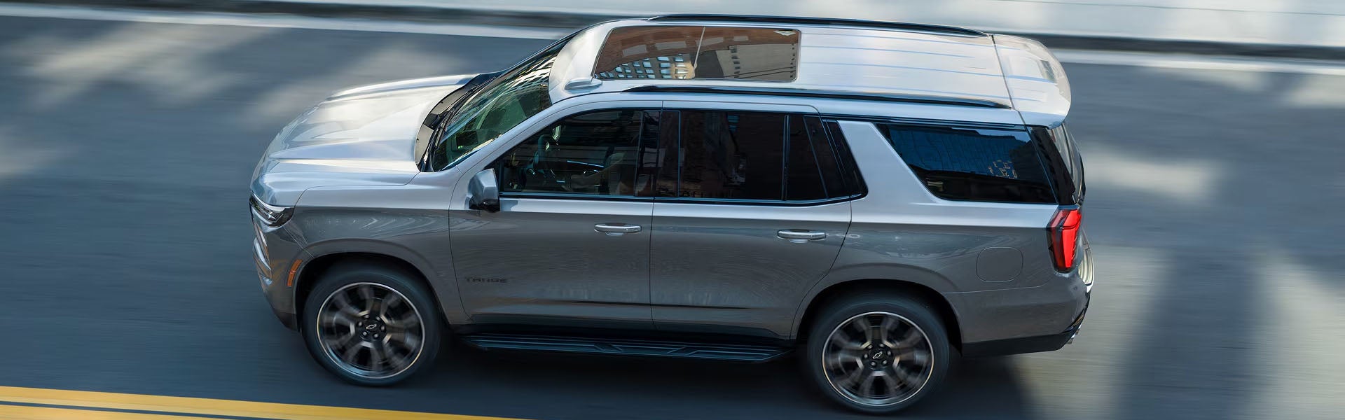 An high-angle side view of a metallic gray Chevrolet Tahoe SUV driving along an asphalt road with a sunroof and tinted windows.